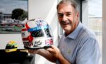 Smiling older man in a light blue striped shirt holds a decorated racing helmet with Union Jack graphics indoors by a window and a photo on the wall behind him.