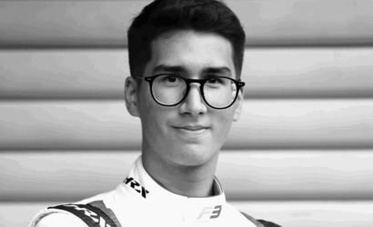 Portrait of a young man with round black glasses, wearing a racing suit, looking at the camera.