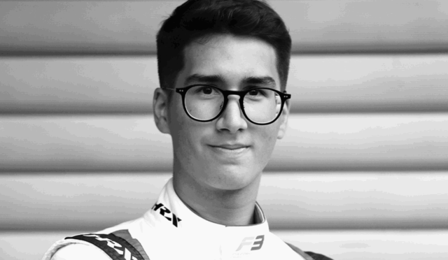 Portrait of a young man with round black glasses, wearing a racing suit, looking at the camera.