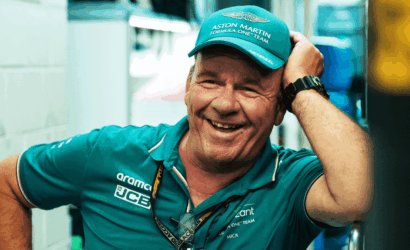Smiling man in teal Aston Martin Formula One Team cap and polo, leaning with hand on his head.