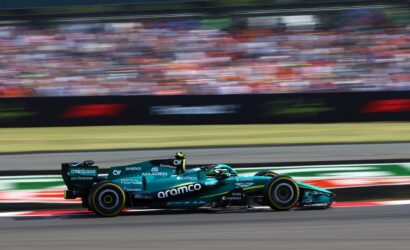 Teal Formula 1 car racing at full speed on a track, with a blurred grandstand in the background.