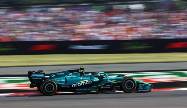 Teal Formula 1 car racing at full speed on a track, with a blurred grandstand in the background.