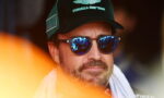 Man wearing a green Aston Martin Formula One Team cap and blue mirrored sunglasses with a white towel around his neck, looking forward.