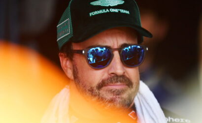 Man wearing a green Aston Martin Formula One Team cap and blue mirrored sunglasses with a white towel around his neck, looking forward.
