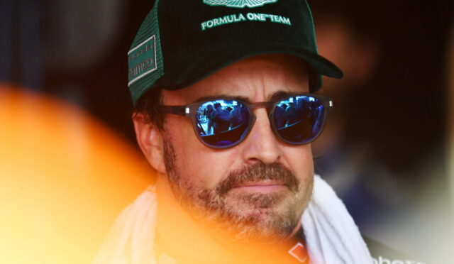 Man wearing a green Aston Martin Formula One Team cap and blue mirrored sunglasses with a white towel around his neck, looking forward.