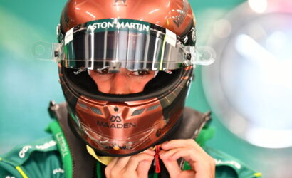 Close-up of a race car driver in a bronze helmet with Aston Martin branding, tightening the visor strap.