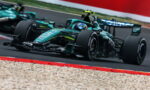 Green Formula 1 race car corners at high speed on a race track with gravel trap and red barriers nearby.
