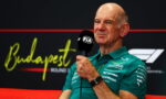 Older man in a teal polo shirt speaks into a black FIA microphone during a press interview, Budapest backdrop behind him.