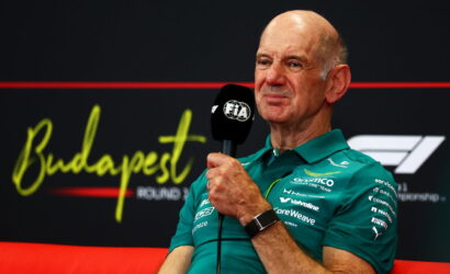 Older man in a teal polo shirt speaks into a black FIA microphone during a press interview, Budapest backdrop behind him.