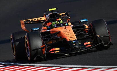 Orange Formula 1 car takes a sharp turn on a race track, neon helmet visible in the cockpit and sponsor logos along the nose and wings