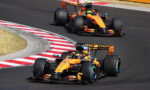 Two orange Formula 1 cars cornering on a race track with red-and-white curbs in the foreground.