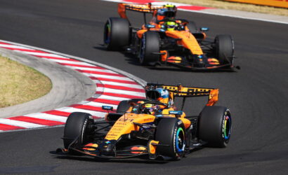 Two orange Formula 1 cars cornering on a race track with red-and-white curbs in the foreground.