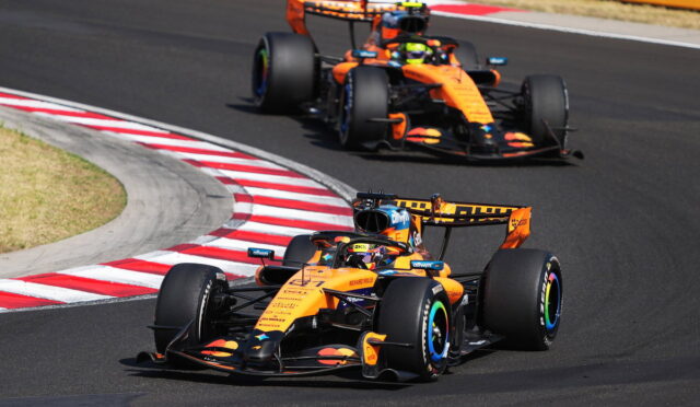 Two orange Formula 1 cars cornering on a race track with red-and-white curbs in the foreground.