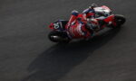 Overhead shot of a Honda racing motorcycle and rider (number 30) leaning into a sharp turn on a dark race track.