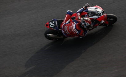 Overhead shot of a Honda racing motorcycle and rider (number 30) leaning into a sharp turn on a dark race track.