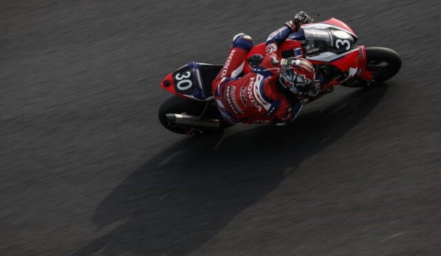 Overhead shot of a Honda racing motorcycle and rider (number 30) leaning into a sharp turn on a dark race track.