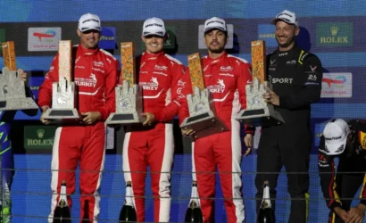 Race car drivers in red uniforms stand on a podium, each holding tall grey trophies with orange accents, Champagne bottles at their feet. Embodies a winner’s celebration backdrop with sponsor logos behind them.