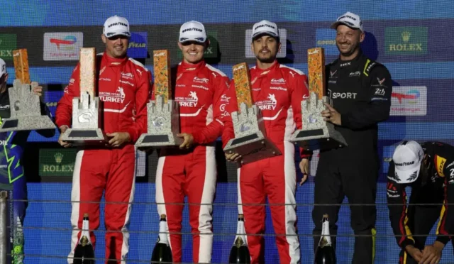 Race car drivers in red uniforms stand on a podium, each holding tall grey trophies with orange accents, Champagne bottles at their feet. Embodies a winner’s celebration backdrop with sponsor logos behind them.