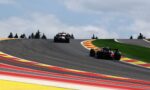 Two Formula 1 cars race around a left-hand curve on a track with bright red, yellow, and white curbs and a green infield under a cloudy sky.