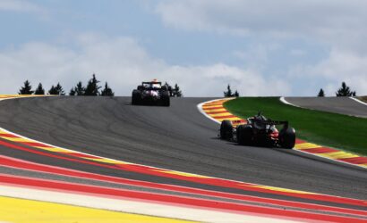 Two Formula 1 cars race around a left-hand curve on a track with bright red, yellow, and white curbs and a green infield under a cloudy sky.