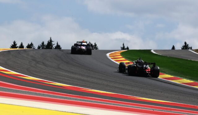 Two Formula 1 cars race around a left-hand curve on a track with bright red, yellow, and white curbs and a green infield under a cloudy sky.