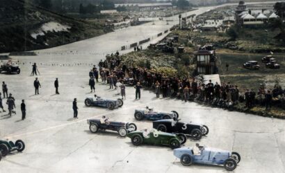 Vintage open-wheel race cars line up at the starting grid as spectators watch along a wide track, 1930s setting.