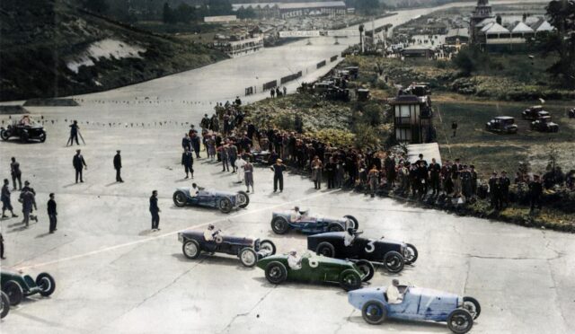 Vintage open-wheel race cars line up at the starting grid as spectators watch along a wide track, 1930s setting.