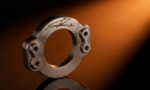 Close-up of a polished metallic circular component with signature engraving and bolt holes, set against an orange gradient background.