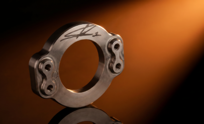 Close-up of a polished metallic circular component with signature engraving and bolt holes, set against an orange gradient background.