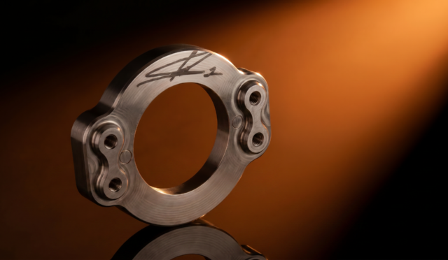 Close-up of a polished metallic circular component with signature engraving and bolt holes, set against an orange gradient background.