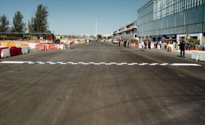 Wide street or runway with a checkered finish line across the foreground, orange barriers, and groups of people along the right beside a glass-fronted building.