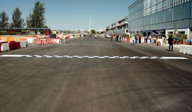 Wide street or runway with a checkered finish line across the foreground, orange barriers, and groups of people along the right beside a glass-fronted building.