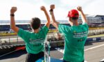 Two people in teal shirts raise their fists in celebration on a race track, with a trophy on the table in front of them.