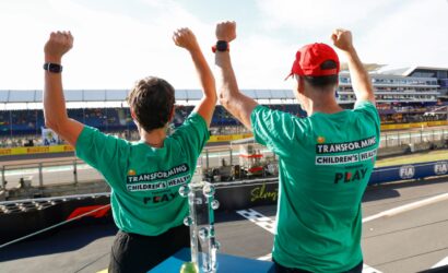 Two people in teal shirts raise their fists in celebration on a race track, with a trophy on the table in front of them.