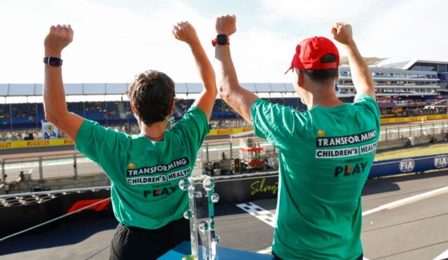 Two people in teal shirts raise their fists in celebration on a race track, with a trophy on the table in front of them.