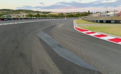 Wide view of a race track curve with red-and-white curbs, safety barriers, and empty spectator fencing under a blue sky