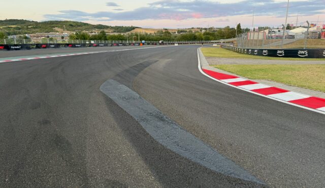 Wide view of a race track curve with red-and-white curbs, safety barriers, and empty spectator fencing under a blue sky