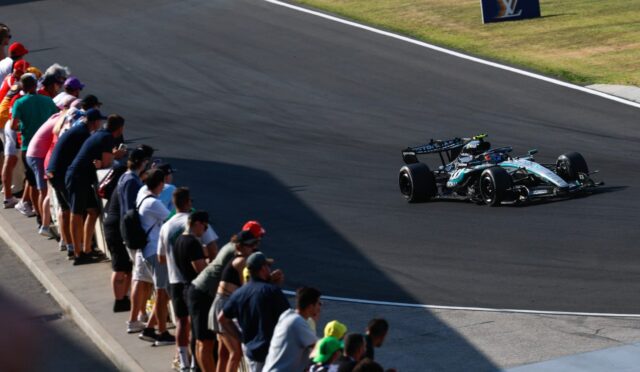 Formula race car speeds along a wide, dark asphalt track while a line of spectators leans over the barrier to watch. A LV sign sits on the grassy infield.