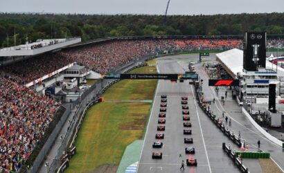 A crowded Formula 1 start line with numerous cars lined up on the grid and a large cheering crowd in the stands.