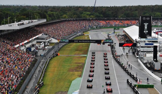 A crowded Formula 1 start line with numerous cars lined up on the grid and a large cheering crowd in the stands.