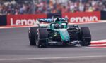 Green Formula 1 car speeding on a race track with blurred crowd and red barriers in the background.