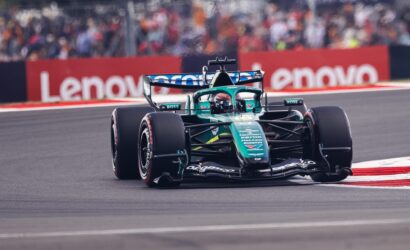Green Formula 1 car speeding on a race track with blurred crowd and red barriers in the background.