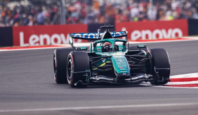 Green Formula 1 car speeding on a race track with blurred crowd and red barriers in the background.