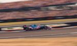 Race car in pink/blue BWT livery speeding along a racetrack with blurred grandstands in the background.