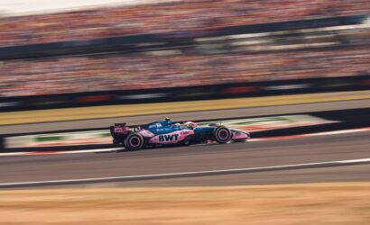 Race car in pink/blue BWT livery speeding along a racetrack with blurred grandstands in the background.