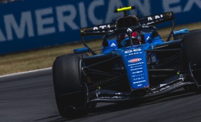 Blue Formula 1 race car taking a corner on a race track, front wing and sponsor logos visible, motion blur on tires in the foreground.