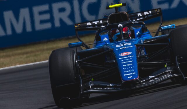 Blue Formula 1 race car taking a corner on a race track, front wing and sponsor logos visible, motion blur on tires in the foreground.