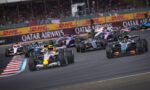 Front row of Formula 1 cars racing on a circuit with a cheering crowd and DHL banners along the barriers.
