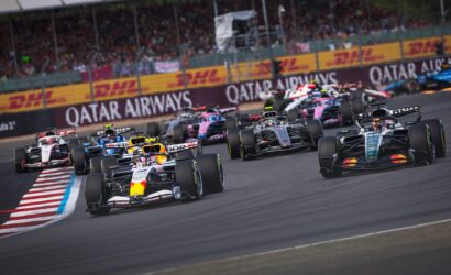 Front row of Formula 1 cars racing on a circuit with a cheering crowd and DHL banners along the barriers.