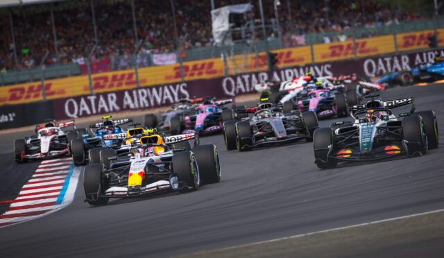 Front row of Formula 1 cars racing on a circuit with a cheering crowd and DHL banners along the barriers.